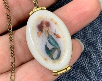 May include: A gold chain necklace with a white oval locket featuring a hand-painted mermaid with a green tail and gold stars. The locket has a hinged opening.