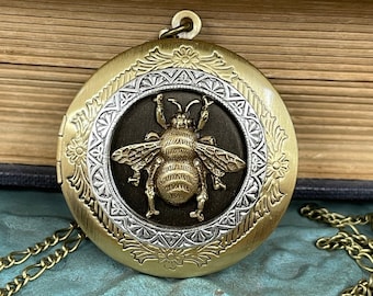 May include: A gold locket necklace with a bee design. The locket has a detailed engraved border and a black background. The bee is gold and has a textured surface.