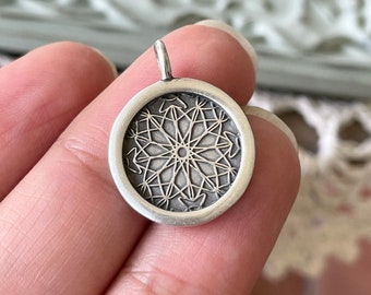 May include: A silver pendant with a detailed geometric design in the center. The pendant is round and has a smooth, polished finish.