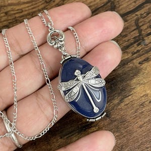 May include: A silver chain necklace with a blue oval pendant featuring a silver dragonfly. The pendant has a hinged opening.