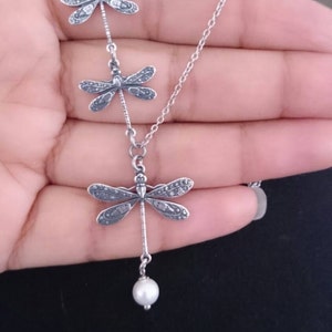 May include: A silver necklace featuring three dragonfly pendants, each with detailed wing patterns. The bottom dragonfly has a dangling white pearl. The necklace is held against a dark background.