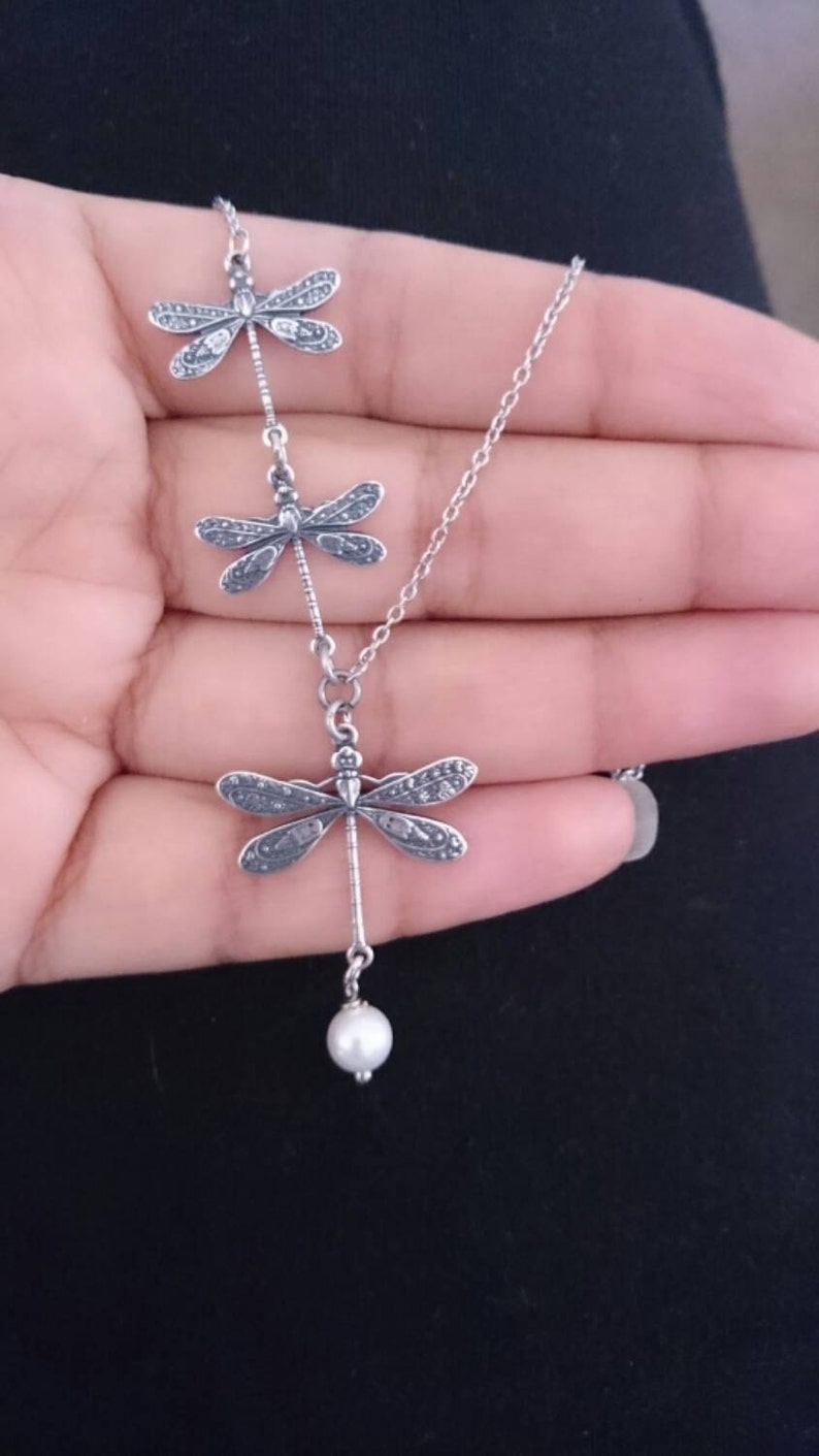 May include: A silver necklace featuring three dragonfly pendants, each with detailed wing patterns. The bottom dragonfly has a dangling white pearl. The necklace is held against a dark background.