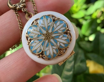 May include: A white porcelain locket necklace with a blue and white floral design. The locket has a hinged opening and a gold-toned chain.