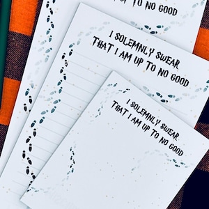 May include: White notepads with the text "I SOLEMNLY SWEAR THAT I AM UP TO NO GOOD" and a green pen are displayed on a black and orange plaid surface. The notepads have a footprint design.