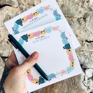 May include: Two white square note pads featuring a border of colorful illustrated cat faces in shades of blue, pink, orange, and black. The top pad displays the text 'a note from JUDY HEINTZ' and the bottom pad reads 'a note from AMANDA KOLKOWSKI'.