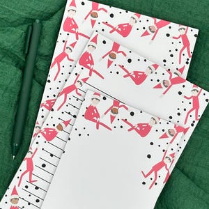 Peut inclure: Une pile de blocs-notes blancs avec un motif d'elfe rouge et des pois noirs. Un stylo vert repose sur un fond de tissu vert. Les blocs-notes ont des lignes noires pour l'écriture.