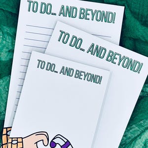 May include: White notepads with the text "TO DO... AND BEYOND!" in green. The notepads feature a cartoon illustration of hands forming a heart shape. A green pen rests on the left side of the image, with a green fabric background.