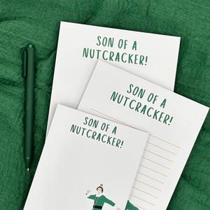 Peut inclure: Bloc-notes blancs avec le texte "SON OF A NUTCRACKER!" en vert. L'un des blocs-notes présente une illustration d'une personne vêtue d'une tenue verte et jaune. Un stylo vert est posé à côté des blocs-notes sur un tissu vert.