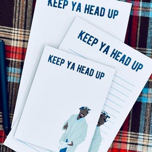 May include: Three white notepads with the text "KEEP YA HEAD UP" in blue. One notepad features an illustration of a person in a light blue coat. A black pen rests on the plaid background.