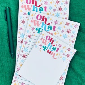 Peut inclure: Trois blocs-notes avec l'inscription "Oh What Fun" en texte coloré, bordés de motifs de flocons de neige et d'étoiles. Un stylo vert est posé à gauche. Le fond est un tissu vert.