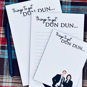 Puede incluir: Tres blocs de notas blancos con el texto "Things to get DUN DUN..." en escritura negra. Un bloc de notas tiene páginas rayadas, y otro presenta un gráfico de dos personas. Un bolígrafo azul oscuro descansa sobre la superficie a cuadros.