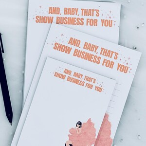 May include: A stack of white notepads with the text "AND, BABY, THAT'S SHOW BUSINESS FOR YOU" in orange. One notepad features a pink illustration of a person sitting on a chair. A black pen rests on the side.