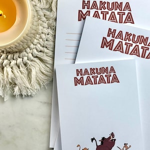 May include: White notepads with the phrase "HAKUNA MATATA" in brown lettering. One notepad features a cartoon illustration of Simba, Pumbaa, and Timon. A lit candle in a white ceramic holder sits on a macrame coaster.