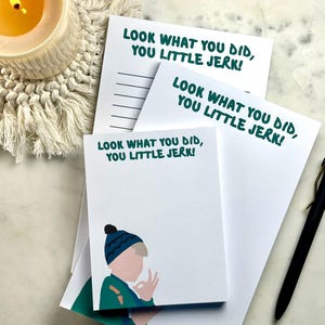 Peut inclure: Bloc-notes et papier blancs avec le texte "LOOK WHAT YOU DID, YOU LITTLE JERK!" en vert. Un bloc-notes présente une illustration d'enfant. Un stylo noir et une bougie allumée sont également visibles.