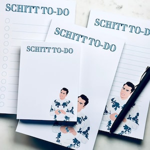 May include: A collection of white notepads with the text "SCHITT TO-DO" in teal. Some pads feature a cartoon illustration of a man in a floral sweater. A black pen rests on one of the pads, ready for use.
