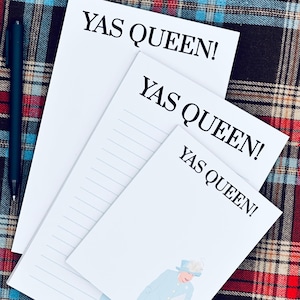May include: Three white notepads with the text "YAS QUEEN!" in black. One notepad features an illustration of a woman in a light blue coat and hat, carrying two black handbags. A dark pen rests on the plaid background.