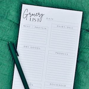Peut inclure: Bloc-notes de liste de courses blanc avec des sections pour la viande/protéines, les produits laitiers/charcuterie, les produits secs, les produits, les surgelés et les articles ménagers. Les mots "Grocery List" sont écrits en cursive. Un stylo vert foncé repose sur le bloc-notes.