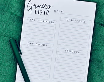 May include: A white grocery list notepad with sections for meat/protein, dairy/deli, dry goods, produce, frozen, and household items. The words "Grocery List" are written in cursive. A dark green pen rests on the notepad, which is on a green fabric surface.
