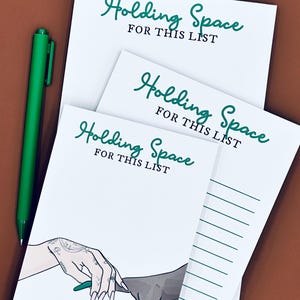 May include: White notepads with the text "Holding Space FOR THIS LIST" in green script. One notepad features an illustration of hands with green fingernails. A green pen rests on the side.