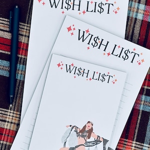 May include: Three white notepads with the text "WISH LIST" in black, accented with red starbursts. One notepad features an illustration of a dancer. A black pen rests on the plaid surface.