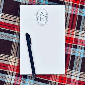 May include: A white notepad with a black pen on a plaid background. The notepad features a geometric design with the letters "A" and "H" inside. The plaid pattern includes red, brown, and blue colors.