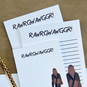 Puede incluir: Blocs de notas blancos con el texto "RAWRGWAWGGGR!" en negro. Un bloc de notas presenta ilustraciones de Chewbacca con un bláster y una tabla de altura. Se ve un bolígrafo dorado con puntos blancos.