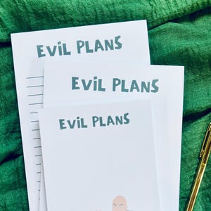 Puede incluir: Tres juegos de bloc de notas blancos con el texto "Evil Plans" impreso en verde en cada uno. El bloc de notas superior tiene papel rayado, el bloc de notas del medio está en blanco y el bloc de notas inferior tiene una ilustración de dibujos animados de un hombre calvo con una camisa verde haciendo un gesto con las manos.