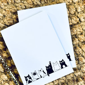 May include: Two white rectangular notepads featuring a row of illustrated black and white cats along the bottom edge. One cat is raising a paw, and another cat is peeking from the side. A gold pen with white polka dots lies to the left.