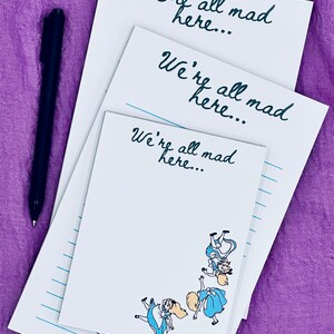 Könnte beinhalten: Drei weiße Notizblöcke mit dem Text "We're all mad here..." in türkisfarbener Schrift. Ein Notizblock zeigt eine Illustration von Alice im Wunderland-Charakteren. Ein schwarzer Stift liegt auf der violetten Oberfläche neben den Notizblöcken.