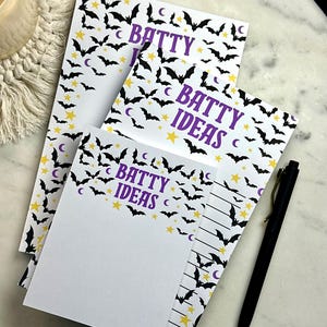 May include: A stack of white notepads with a Halloween theme. The pads feature a pattern of black bats, yellow stars, and purple text that reads "BATTY IDEAS". A black pen rests on the side.
