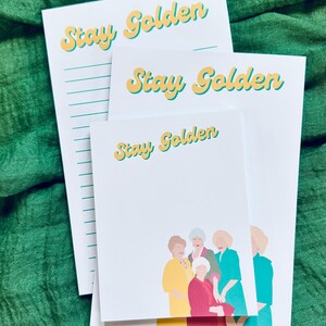 K&ouml;nnte beinhalten: Wei&szlig;e Notizbl&ouml;cke mit dem Text "Stay Golden" in Gelb und T&uuml;rkis. Ein Notizblock zeigt eine Gruppenillustration in Gelb, Rot und T&uuml;rkis. Die Notizbl&ouml;cke sind auf einer strukturierten gr&uuml;nen Oberfl&auml;che gestapelt.