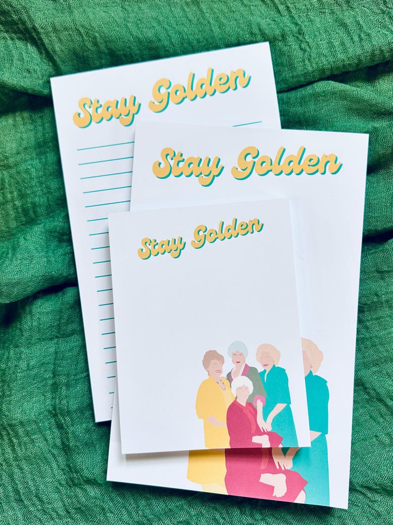 K&ouml;nnte beinhalten: Wei&szlig;e Notizbl&ouml;cke mit dem Text "Stay Golden" in Gelb und T&uuml;rkis. Ein Notizblock zeigt eine Gruppenillustration in Gelb, Rot und T&uuml;rkis. Die Notizbl&ouml;cke sind auf einer strukturierten gr&uuml;nen Oberfl&auml;che gestapelt.