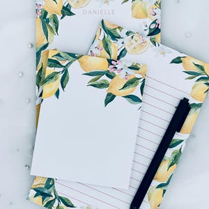 May include: A collection of stationery featuring a lemon theme. The set includes notepads and a lined pad, all adorned with watercolor illustrations of lemons, leaves, and blossoms. The name "DANIELLE" is printed on one notepad. A black pen rests on the lined pad.
