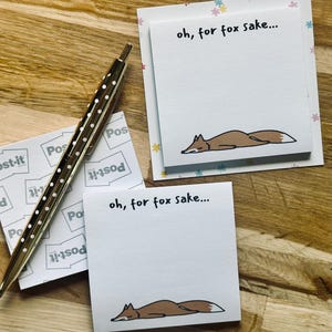 May include: Two square white sticky note pads featuring a brown fox illustration and the text "oh, for fox sake..." at the top, accompanied by a metallic pen with a white polka dot pattern on the barrel, resting on a light wood surface.