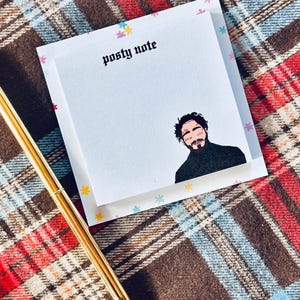 May include: A white sticky note pad with a colorful floral border. The text "Posty Note" is printed in black at the top of the pad. A black and white illustration of a man with a beard is printed on the pad.