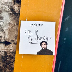 May include: A white sticky note with the text "posty note" at the top. The note says "pick up dry cleaning" in black handwriting. A cartoon illustration of a man with a beard is at the bottom of the note.