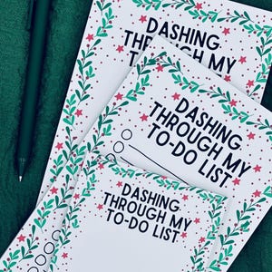 Peut inclure: Bloc-notes blancs avec des motifs de Noël verts et rouges, avec le texte "Dashing Through My To-Do List". Un stylo vert foncé repose sur un tissu vert.