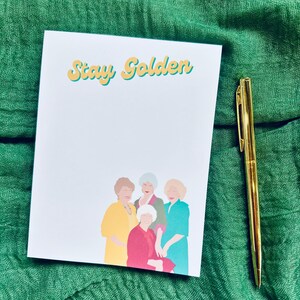 K&ouml;nnte beinhalten: Ein wei&szlig;er Notizblock mit den Worten "Stay Golden" in gelben und blauen Buchstaben. Der Notizblock zeigt eine Illustration von vier Frauen in farbenfrohen Outfits. Ein goldener Stift liegt neben dem Notizblock auf einem gr&uuml;nen Stoff.
