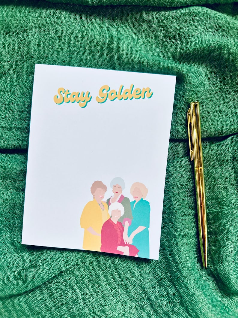 K&ouml;nnte beinhalten: Ein wei&szlig;er Notizblock mit den Worten "Stay Golden" in gelben und blauen Buchstaben. Der Notizblock zeigt eine Illustration von vier Frauen in farbenfrohen Outfits. Ein goldener Stift liegt neben dem Notizblock auf einem gr&uuml;nen Stoff.