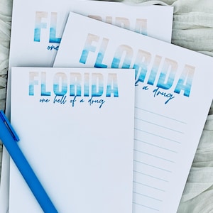 Puede incluir: Blocs de notas y papel rayado blancos con la palabra "FLORIDA" en letras de acuarela azul y beige. La frase "one hell of a drug" está debajo de la palabra "FLORIDA". Un bolígrafo azul descansa sobre el bloc de notas.