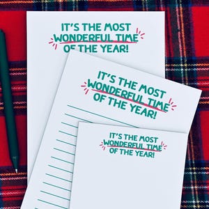 Peut inclure: Trois blocs-notes blancs avec le texte "IT'S THE MOST WONDERFUL TIME OF THE YEAR!" en vert et rouge. Les blocs-notes sont sur un fond à carreaux rouge et vert, avec un stylo vert sur le côté gauche. Le bloc-notes du bas a une ligne de personnages de dessins animés.