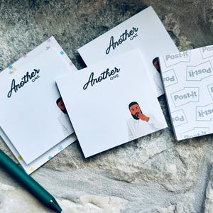 Op de afbeelding: Witte vierkante plaknotities met de tekst "Another One" in een scriptlettertype en een illustratie van een man met een baard. Een groene pen staat links. Een Post-it notitieblok staat rechts.