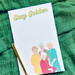 K&ouml;nnte beinhalten: Ein wei&szlig;er Notizblock mit dem Text "Stay Golden" in gelben und t&uuml;rkisfarbenen Buchstaben. Der Notizblock zeigt eine farbenfrohe Illustration von vier Frauen. Ein goldener Stift liegt auf dem Notizblock vor einem gr&uuml;nen Stoffhintergrund.