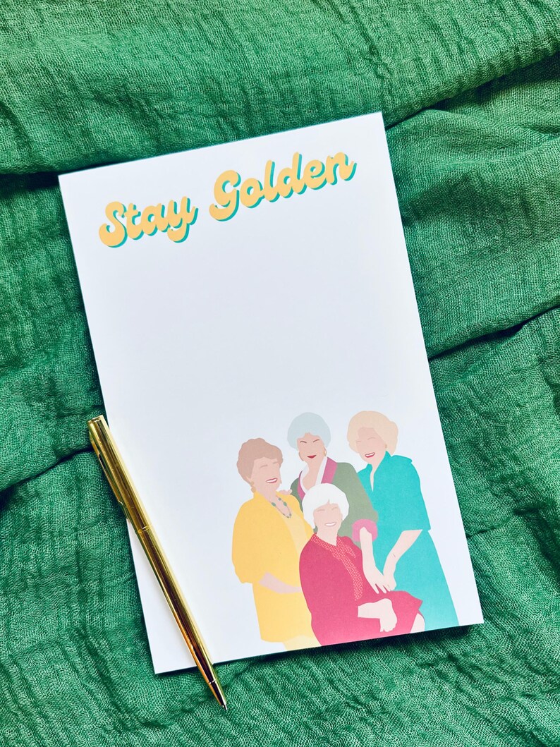 K&ouml;nnte beinhalten: Ein wei&szlig;er Notizblock mit dem Text "Stay Golden" in gelben und t&uuml;rkisfarbenen Buchstaben. Der Notizblock zeigt eine farbenfrohe Illustration von vier Frauen. Ein goldener Stift liegt auf dem Notizblock vor einem gr&uuml;nen Stoffhintergrund.
