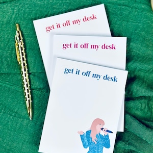 May include: Three white notepads with the text "get it off my desk" in pink, blue, and blue lettering. The bottom notepad features a cartoon illustration of a woman with pink hair wearing a blue sparkly dress and holding a microphone.