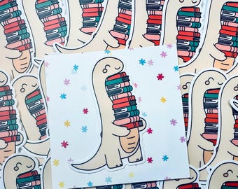 Puede incluir: Una pegatina de un dinosaurio de dibujos animados sosteniendo una pila de libros coloridos. El dinosaurio es beige con la nariz rosa y una expresión feliz. El fondo es blanco con un patrón de pequeñas flores rosas, azules y amarillas.