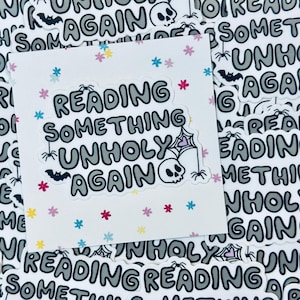 Può includere: Un adesivo bianco con un design nero e grigio. L'adesivo dice "Reading Something Unholy Again" con una ragnatela, un ragno, un pipistrello e un teschio. L'adesivo è circondato da uno sfondo bianco con piccoli fiori rosa, blu e gialli.