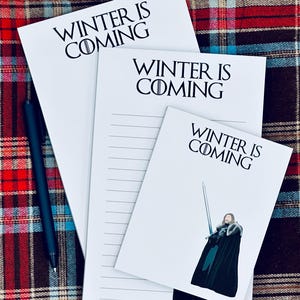 Puede incluir: Tres blocs de notas blancos con el texto "WINTER IS COMING" en negro. Un bloc de notas tiene papel rayado y otro presenta una ilustración de personaje. Un bolígrafo azul oscuro descansa sobre una superficie a cuadros.