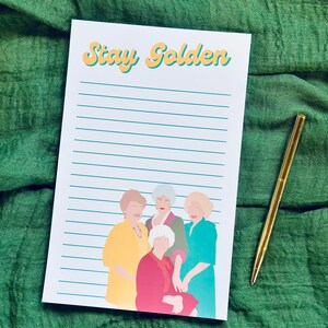 K&ouml;nnte beinhalten: Ein Notizblock mit dem Text "Stay Golden" in gelber und orangefarbener Schrift, mit einer Illustration von vier Frauen in farbenfrohen Outfits. Ein goldener Stift liegt auf einem gr&uuml;nen Stoffhintergrund.