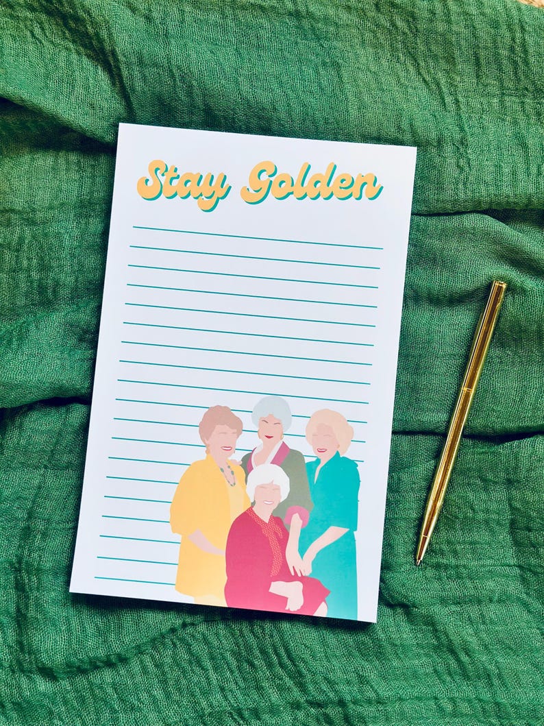 K&ouml;nnte beinhalten: Ein Notizblock mit dem Text "Stay Golden" in gelber und orangefarbener Schrift, mit einer Illustration von vier Frauen in farbenfrohen Outfits. Ein goldener Stift liegt auf einem gr&uuml;nen Stoffhintergrund.